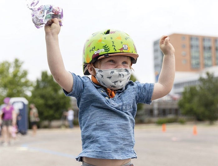 Kid with yellow helmet and a face mask raising both hands in the air outside