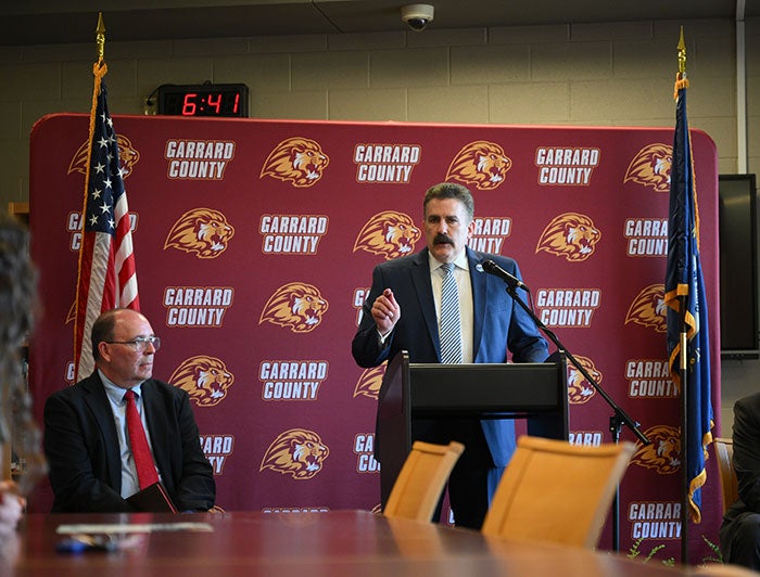 Man standing on podium and speaking through microphone. Behind him is the American flag and a poster board featuring the Gerrard County Lions High School Logo and name. To the right of man on the podium is another man sitting down in a table listening.