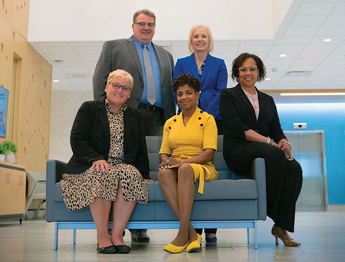 Five people dressed professionally, three of them sitting on a couch while the other two are standing. They are all posing for a picture.