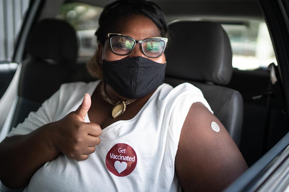Supporting Public Health. A woman wearing a mask with a Get Vaccinated sticker on her shirt shows her vaccine location Band-Aid.