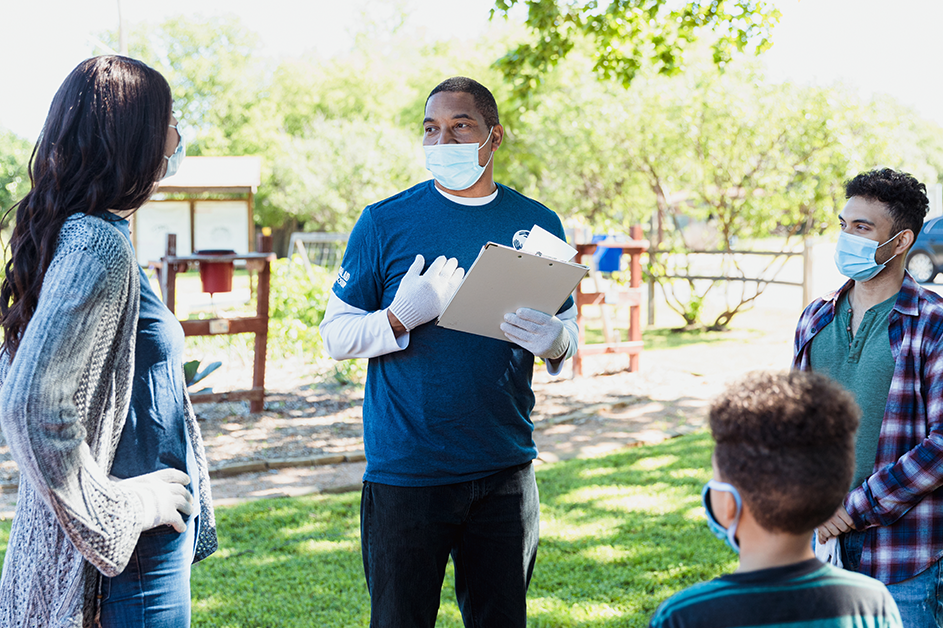 Benefits to Communities. A clinician wearing a mask talks to three community members wearing masks in a park.