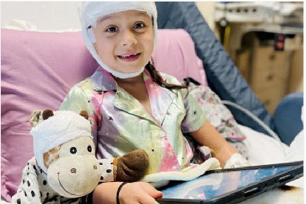 Telling the Hospital Story. A child with head wrapped in gauze and bandages sits upright in a hospital bed holding a tablet computer.