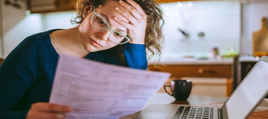 A woman with glasses and a blue sweater examines a bill as she sits in her kitchen. She holds her forehead in her hand.